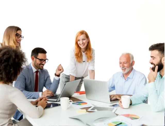 Pessoas de diversas etnias sorrindo à mesa de trabalho, em reunião. Laptops e papéis com gráficos em cima da mesa branca. Algumas canecas brancas. Fundo branco.